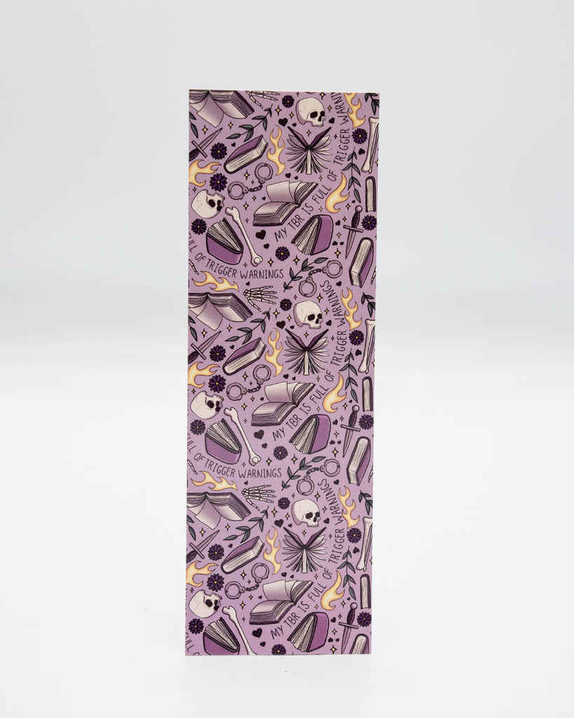 Front view of the My TBR is Full of Trigger Warnings Bookmark from Rebel Romance, featuring a repeat pattern of skulls, books, flames, and gothic florals on a purple background. Ideal for readers of dark romance, fantasy smut, and alternative fiction with a rebellious twist.
