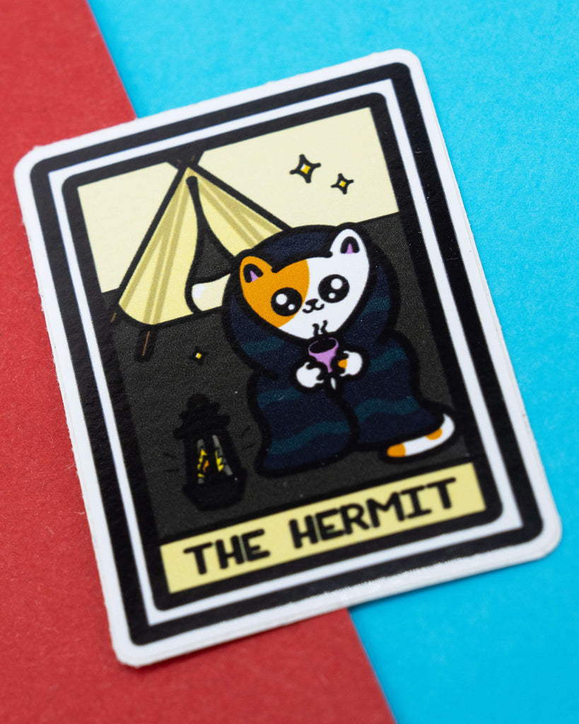 The Hermit Tarot Cat Sticker by Innabox, available at Rebel Romance. Features a cartoon calico cat cloaked in dark robes holding a cup of tea, beside a lantern and tent under a night sky. Glossy waterproof vinyl sticker shown on a colourful red and blue background.
