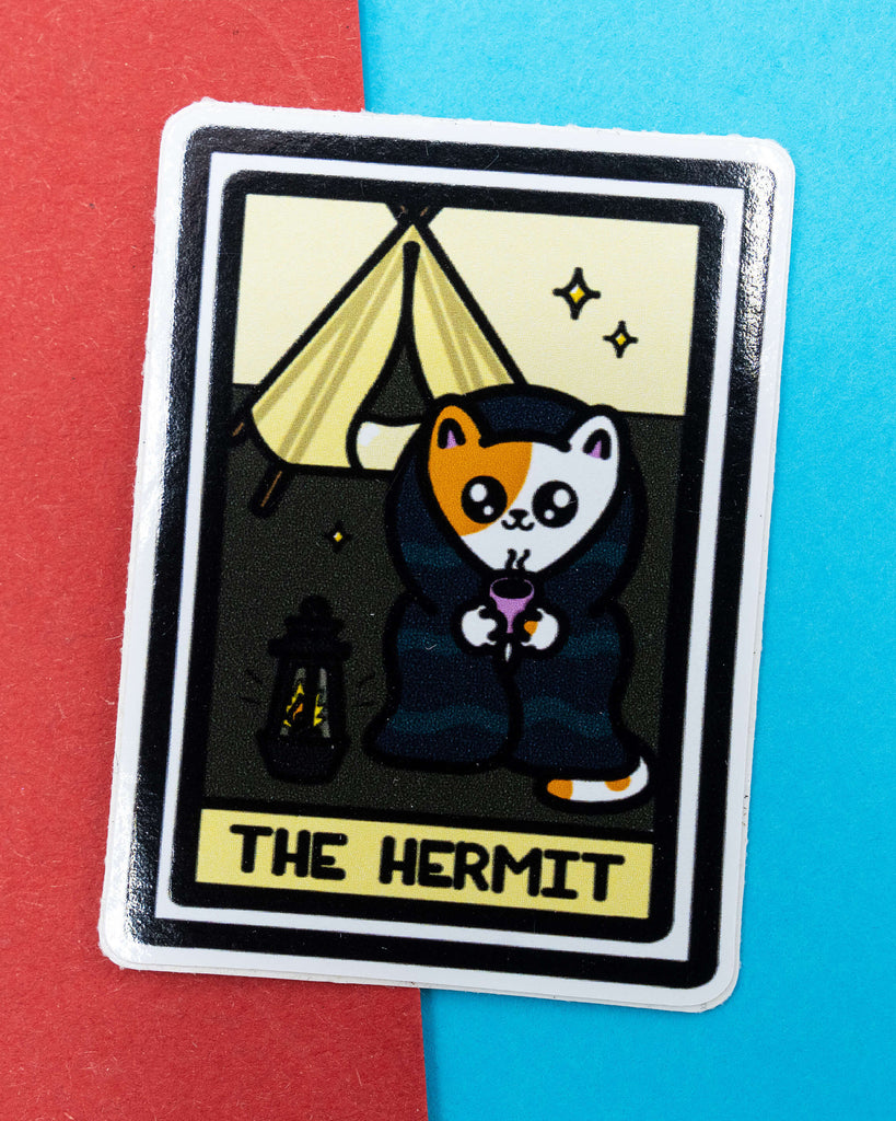Gloss vinyl sticker featuring The Hermit Tarot Card Cat design by Innabox, sold by Rebel Romance. The sticker shows a cute calico cat wrapped in a cloak, holding a steaming mug outside a tent, with a lantern nearby. Ideal for introverts and tarot lovers. Photographed on a red and blue background.