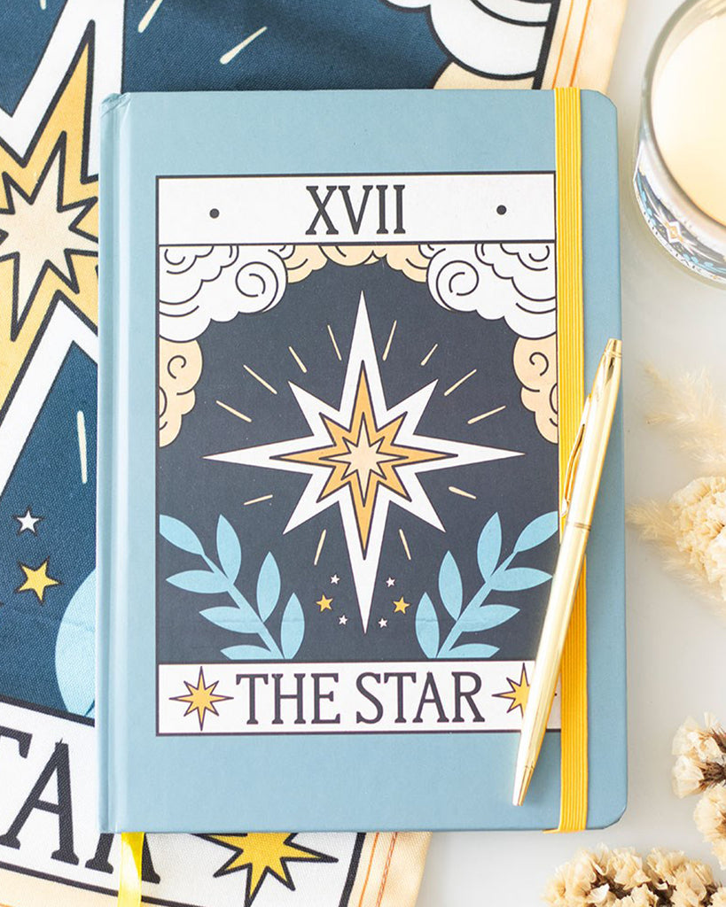 A pastel blue A5 notebook with a vintage-style ‘The Star’ tarot card cover design in orange, white, and blue with celestial symbols. Features a yellow elastic strap and matching ribbon marker.