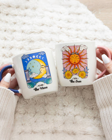 Close-up of a person holding both the Sun and Moon mugs, with white sweater sleeves visible. The detailed tarot-style artwork on each mug is clearly shown, with soft lighting and a textured blanket in the background.
