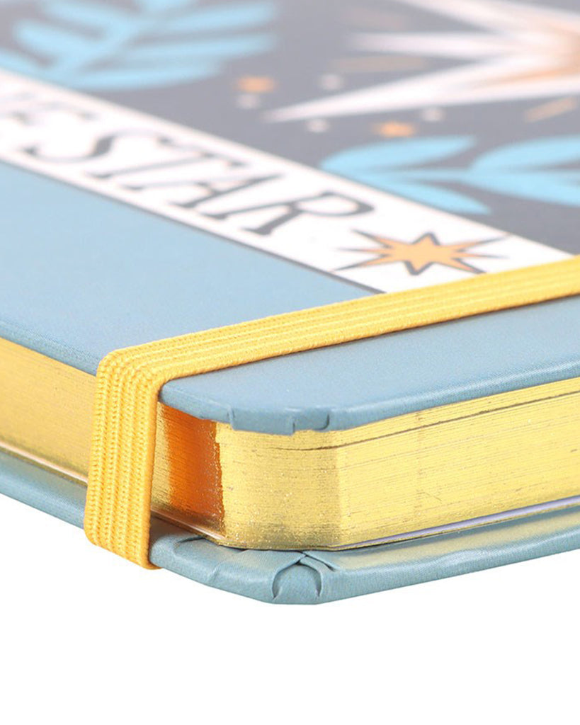 Close-up of the corner of the ‘The Star’ tarot notebook showing gold foil edges and yellow elastic strap against a pastel blue cover.