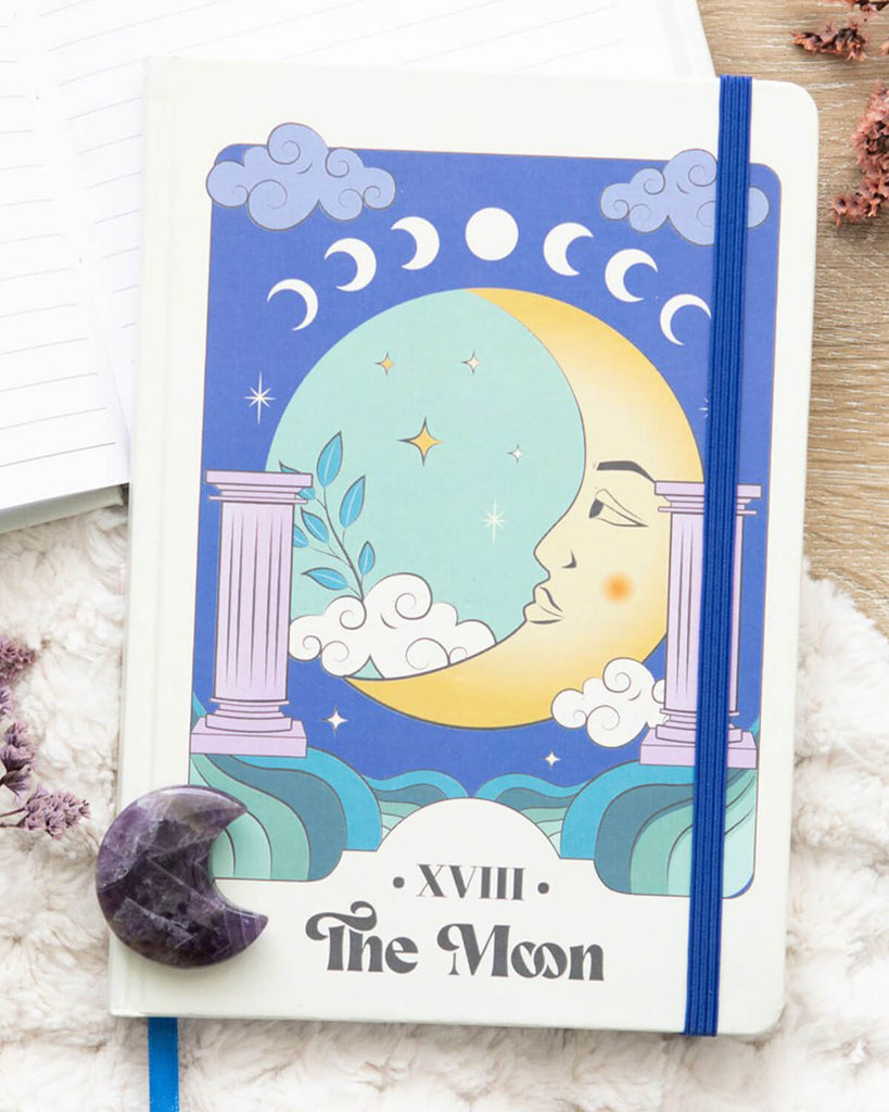 Flat lay of The Moon Celestial A5 Notebook – Tarot-inspired notebook with a crescent moon face, pastel columns, teal sky, and moon phases along the top. “XVIII The Moon” is written at the bottom in bold, vintage-style font.