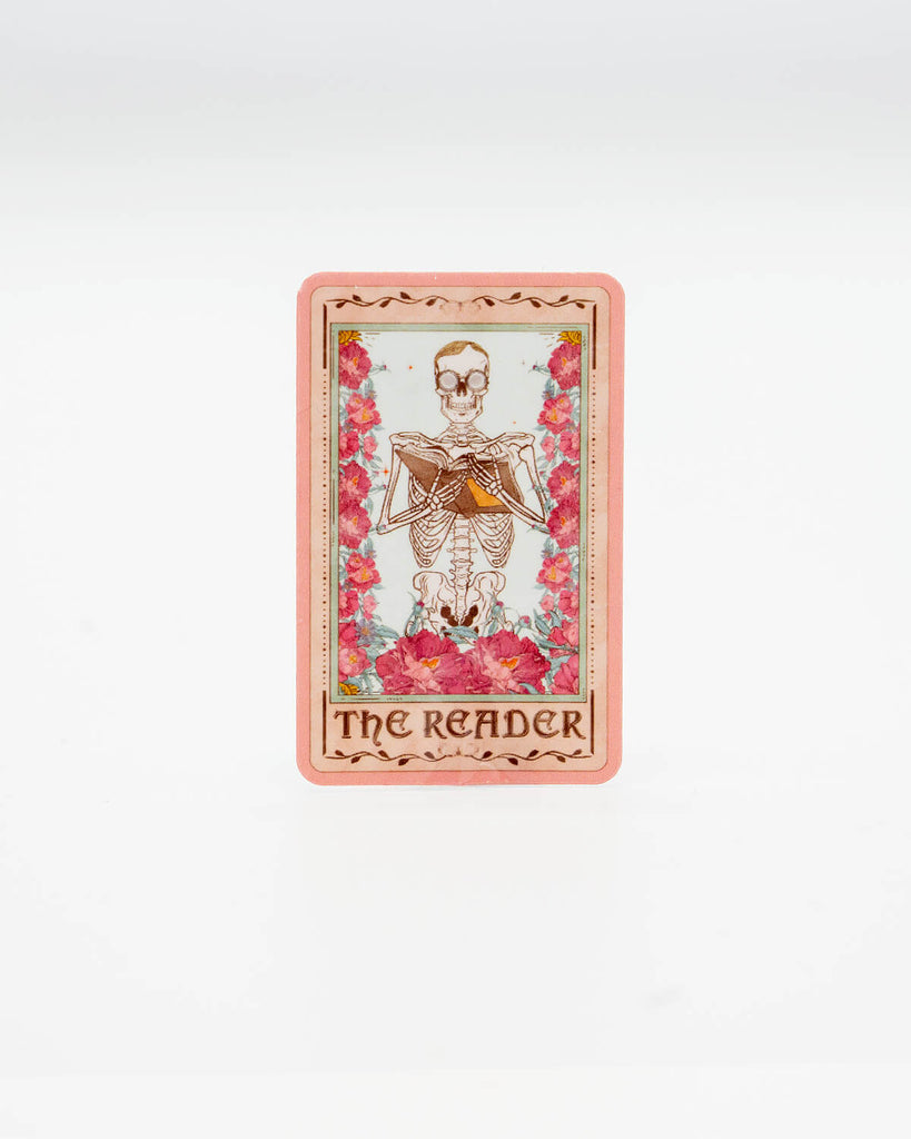 Die-cut vinyl sticker styled like a tarot card, featuring a skeleton reading a book surrounded by pink flowers. The bottom reads “The Reader” in gothic lettering. A mystical, bookish design perfect for fans of dark academia and tarot aesthetics. Created by Rebel Romance, a small UK brand known for alternative style, slow fashion, and quirky millennial-friendly gifts.
