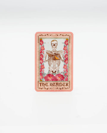 Die-cut vinyl sticker styled like a tarot card, featuring a skeleton reading a book surrounded by pink flowers. The bottom reads “The Reader” in gothic lettering. A mystical, bookish design perfect for fans of dark academia and tarot aesthetics. Created by Rebel Romance, a small UK brand known for alternative style, slow fashion, and quirky millennial-friendly gifts.