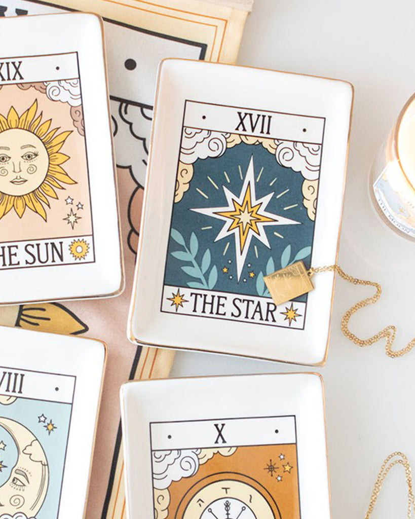 A flat lay of Rebel Romance tarot-inspired ceramic trinket dishes featuring The Star, The Sun and other designs. The Star dish with gold edging is in focus, styled with a gold necklace and mystical-themed accessories, perfect for jewellery and small treasures.