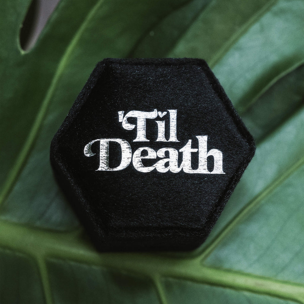 Close-up of a black hexagonal velvet ring box with “’Til Death” embroidered in striking white script, placed on a large green leaf for a dramatic effect.