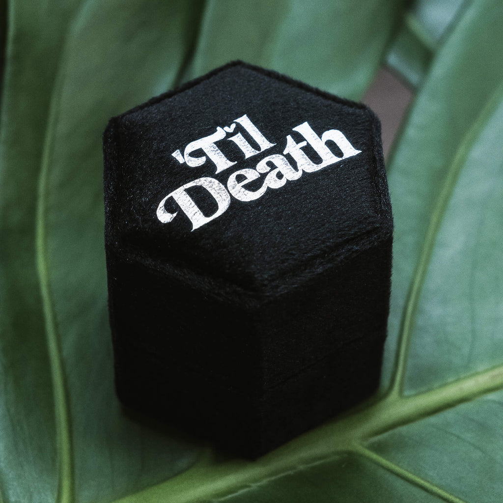 A black hexagonal velvet ring box with “’Til Death” elegantly embroidered in white script on the lid, set against a leafy green background.