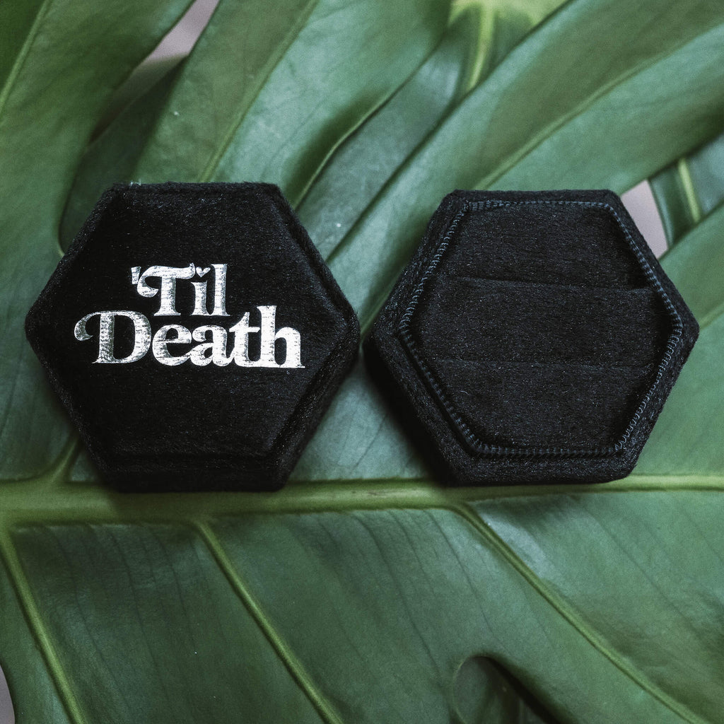 Closed black hexagonal velvet ring box showing “’Til Death” in bold white lettering, alongside an identical empty box, placed on a vibrant green leaf.