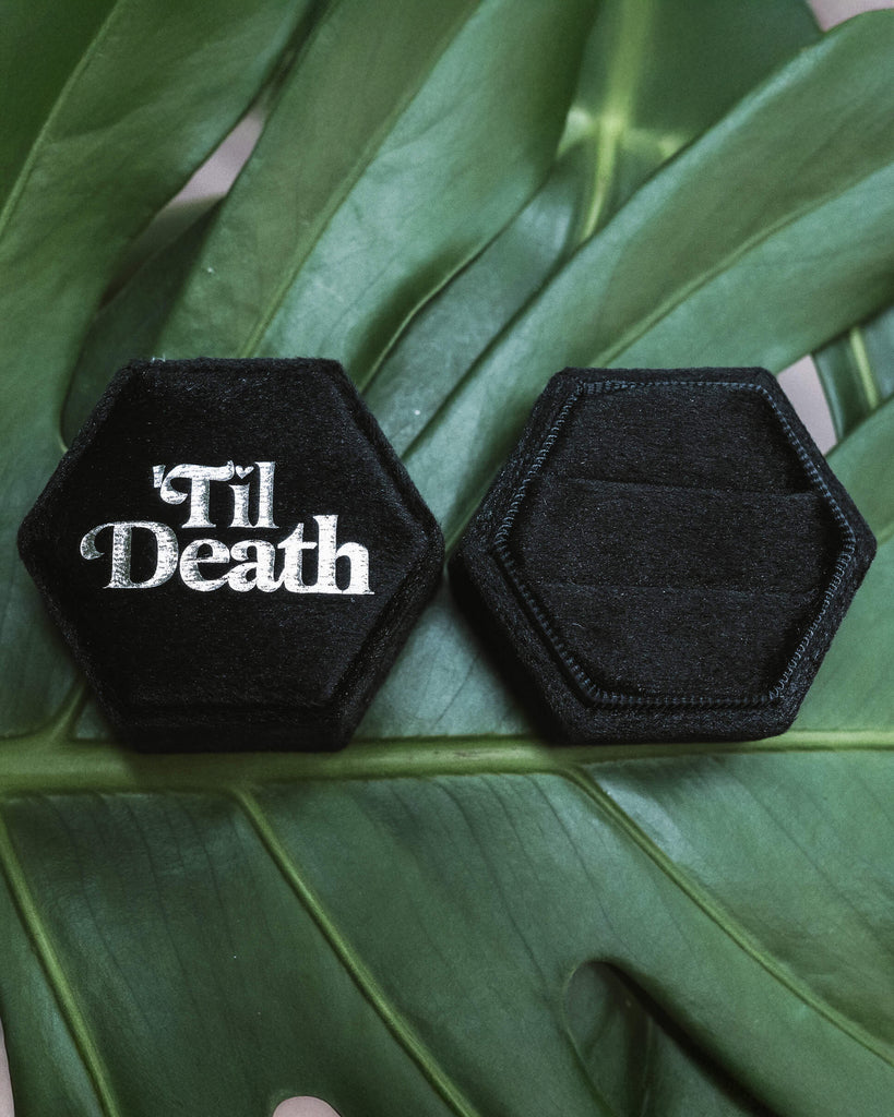 Open black hexagonal velvet ring box showing the top half reading “’Til Death” in bold white lettering, alongside an the other half of an empty box with two slots for rings, placed on a vibrant green leaf.