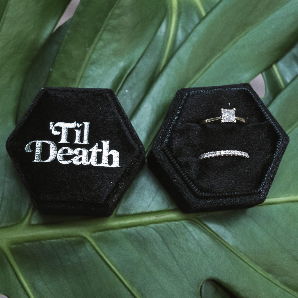 Open black hexagonal velvet ring box featuring “’Til Death” embroidered in white on the lid and two ring slots holding a solitaire engagement ring and a diamond eternity band, displayed on a leafy backdrop.