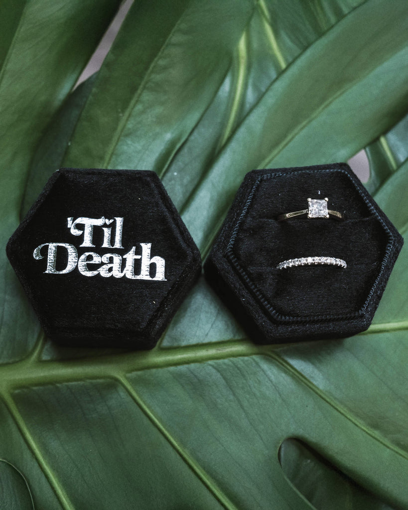Open black hexagonal velvet ring box featuring “’Til Death” embroidered in white on the lid and two ring slots holding a solitaire engagement ring and a diamond eternity band, displayed on a leafy backdrop.