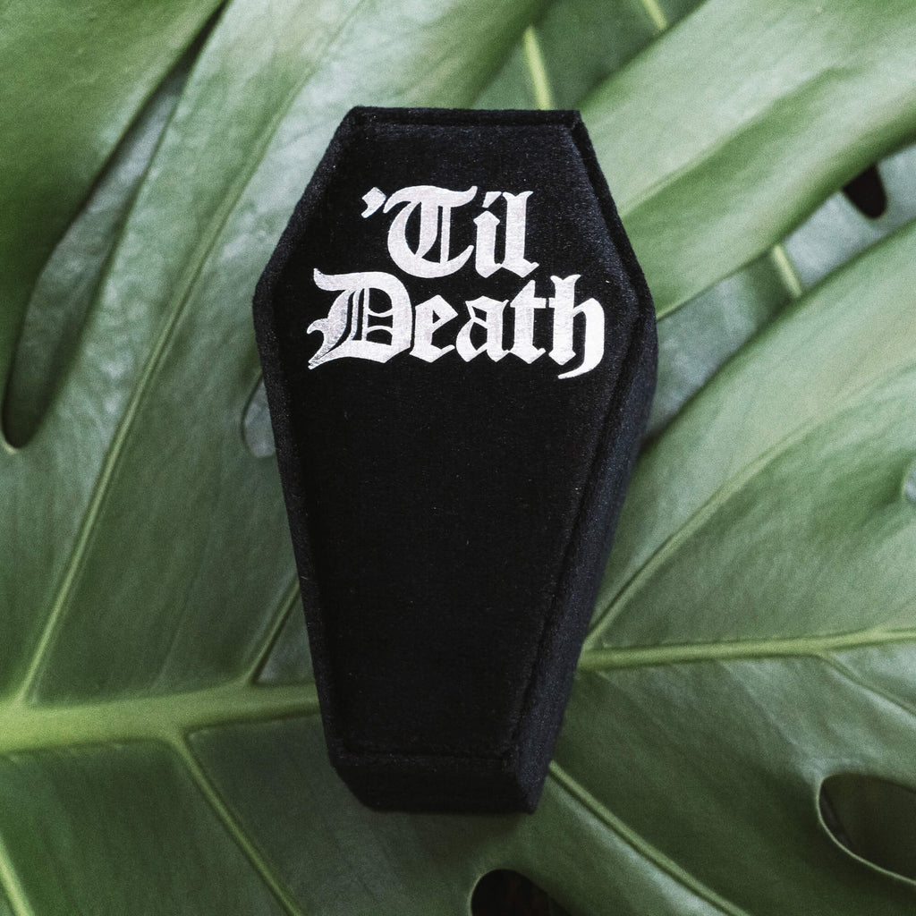 Coffin Shaped Ring Box - Full Display
A black velvet coffin-shaped ring box displayed closed, showcasing the “Til Death” gothic text, and open next to it, revealing the interior slots. Set against a tropical green leaf backdrop for a bold contrast.
