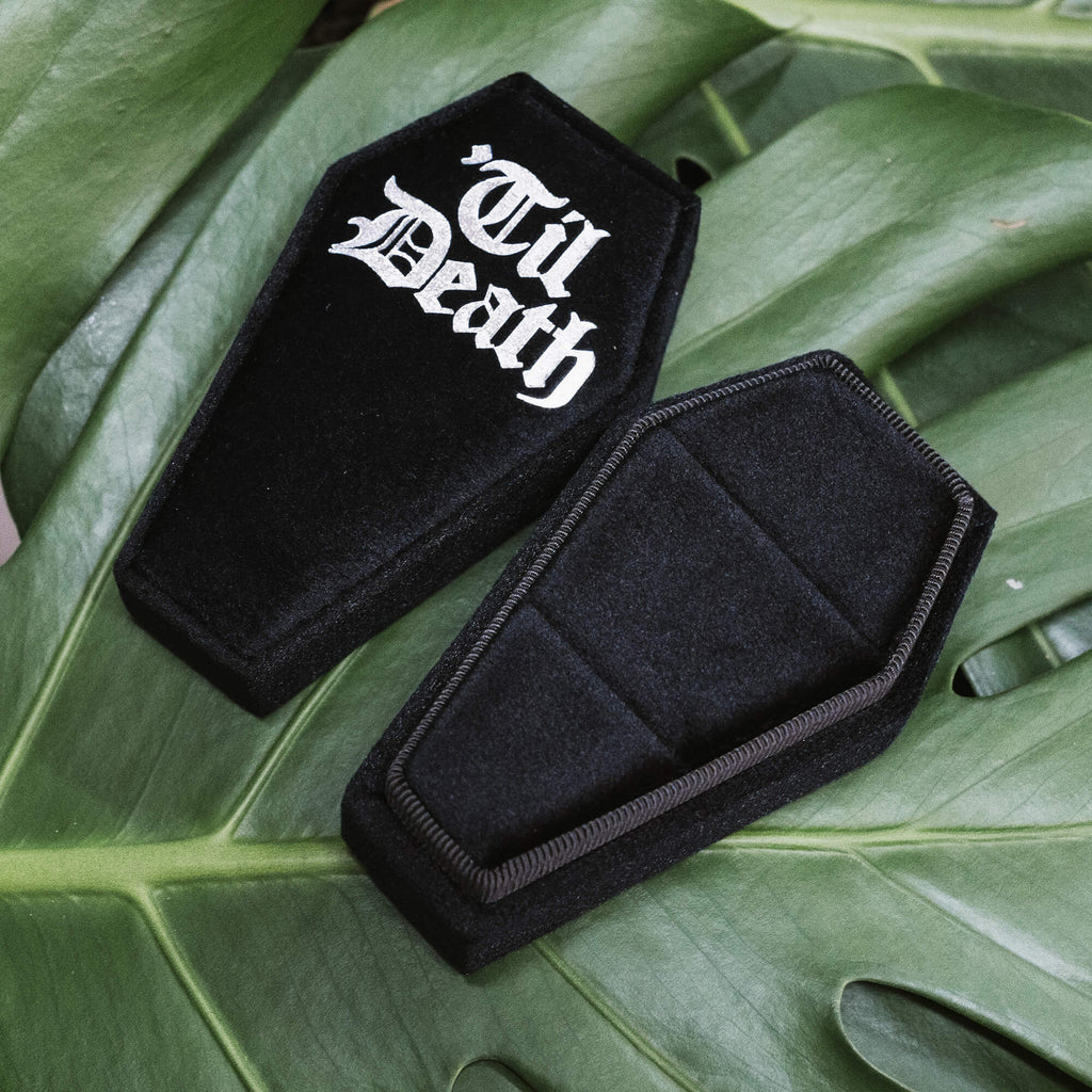 Coffin Shaped Ring Box - Til Death 
A black velvet coffin-shaped ring box with “Til Death” printed in gothic white font on the lid. Set against lush green leaves for a dramatic gothic aesthetic.