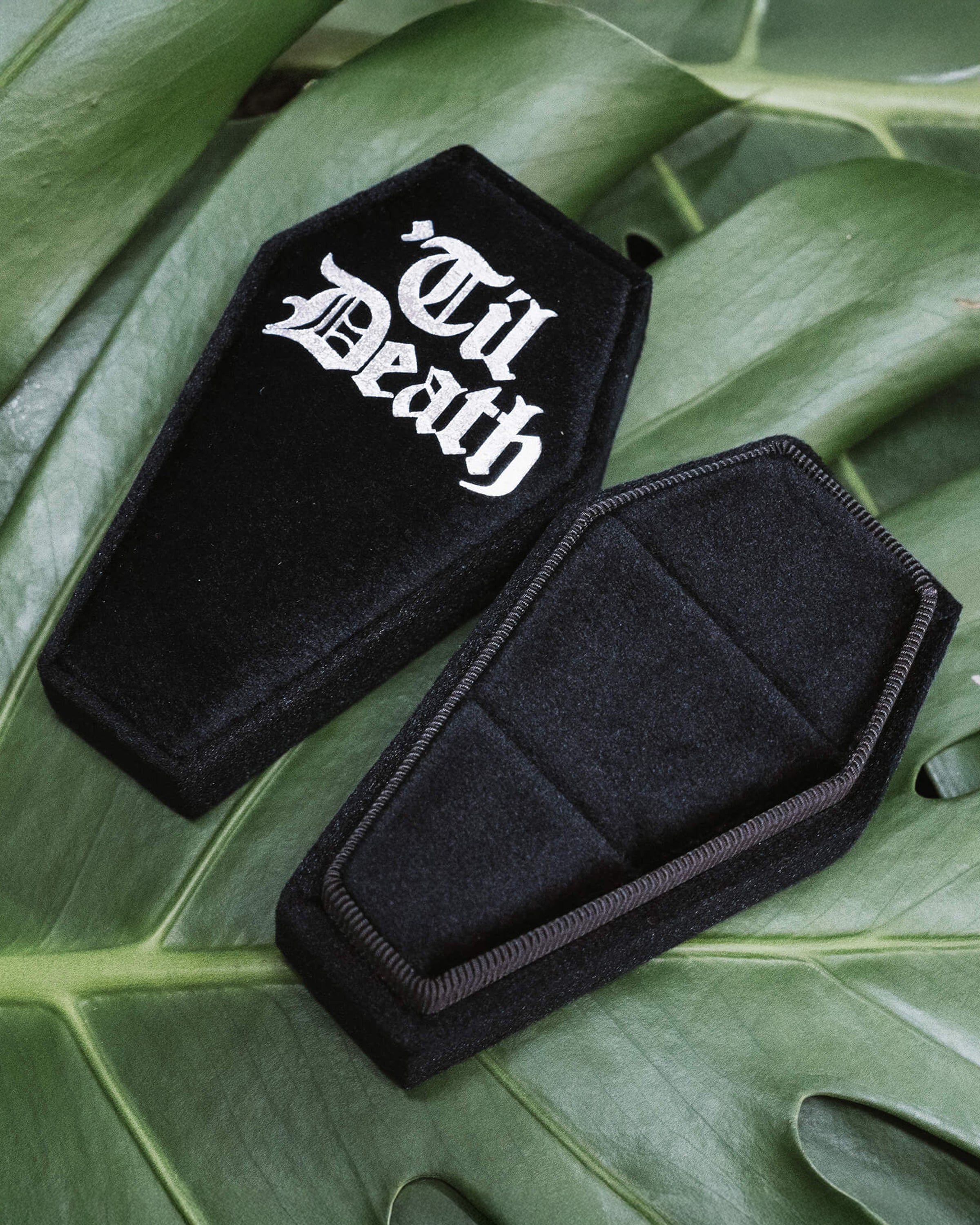 Coffin Shaped Ring Box - Til Death 
A black velvet coffin-shaped ring box with “Til Death” printed in gothic white font on the lid. Set against lush green leaves for a dramatic gothic aesthetic.