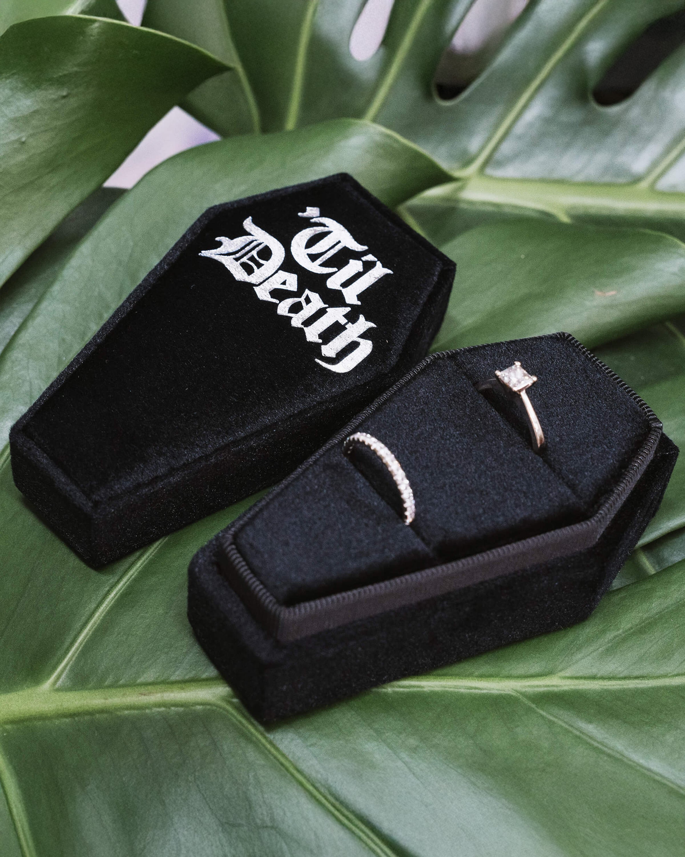 Coffin Shaped Ring Box - Til Death with Rings
A black velvet coffin-shaped ring box featuring two wedding rings—a diamond engagement ring and a band. “Til Death” is printed in gothic white font on the lid, styled against a green leaf background.