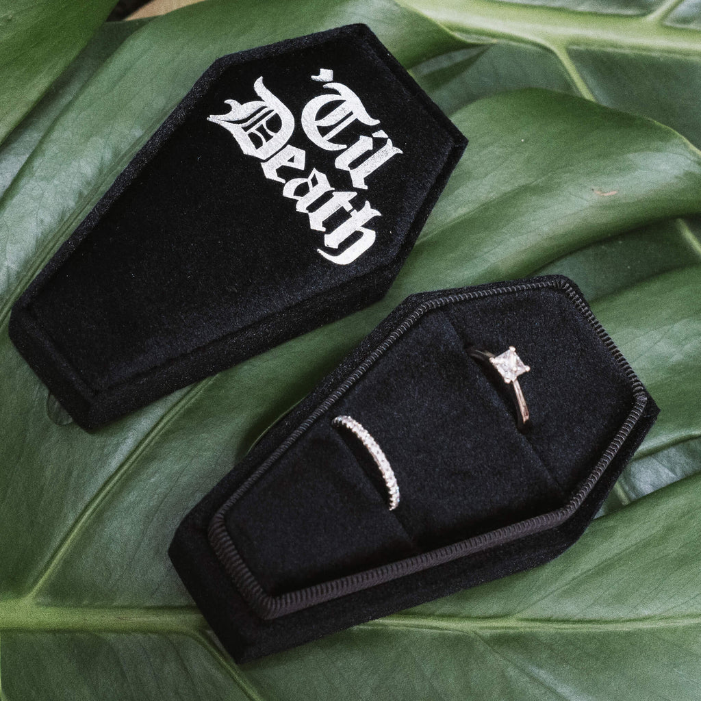 Coffin Shaped Ring Box - Tilted View
A tilted view of a black velvet coffin-shaped ring box, showcasing the gothic white “Til Death” text prominently on the lid, surrounded by lush green leaves.