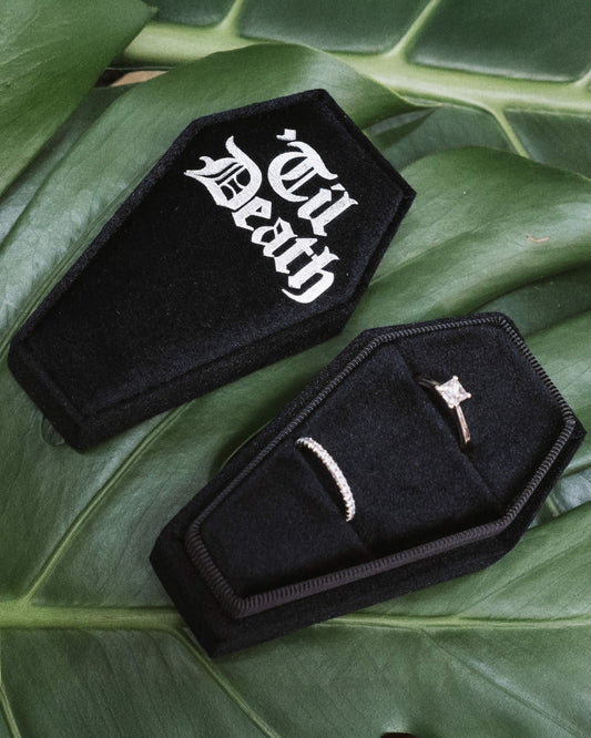 Coffin Shaped Ring Box - Tilted View
A tilted view of a black velvet coffin-shaped ring box, showcasing the gothic white “Til Death” text prominently on the lid, surrounded by lush green leaves.