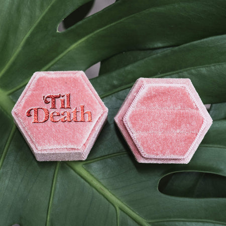 Two pink hexagonal velvet ring boxes side by side, one featuring the phrase “’Til Death” embroidered in red, both displayed on a green monstera leaf.