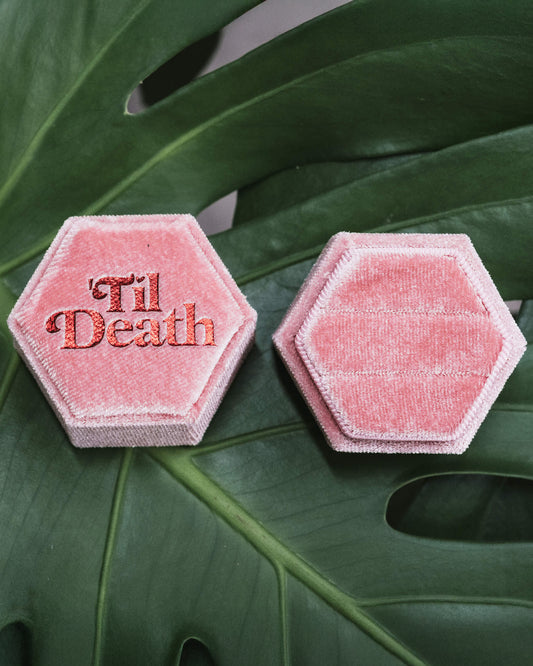 Two pink hexagonal velvet ring boxes side by side, one featuring the phrase “’Til Death” embroidered in red, both displayed on a green monstera leaf.