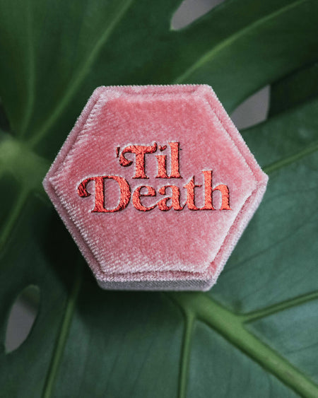 A pink hexagonal velvet ring box with the phrase “’Til Death” embroidered in bold, shiny red letters on top, resting on a green monstera leaf background.