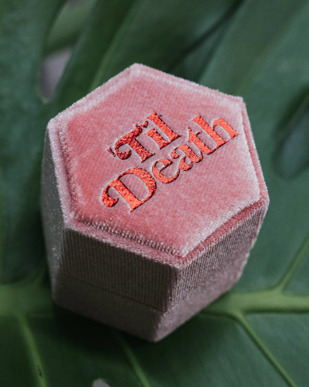 Close-up of a pink hexagonal velvet ring box with “’Til Death” in striking red embroidery, set against a lush green monstera leaf.