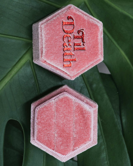 Two open pink hexagonal velvet ring boxes on a green monstera leaf, one showing “’Til Death” embroidered in red and the other holding two elegant silver rings.