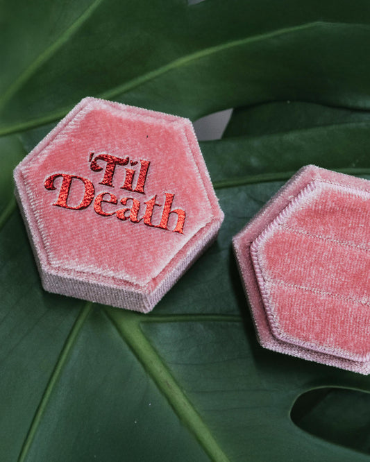 Two open pink hexagonal velvet ring boxes on a green monstera leaf, one showing “’Til Death” embroidered in red and the other holding two elegant silver rings.