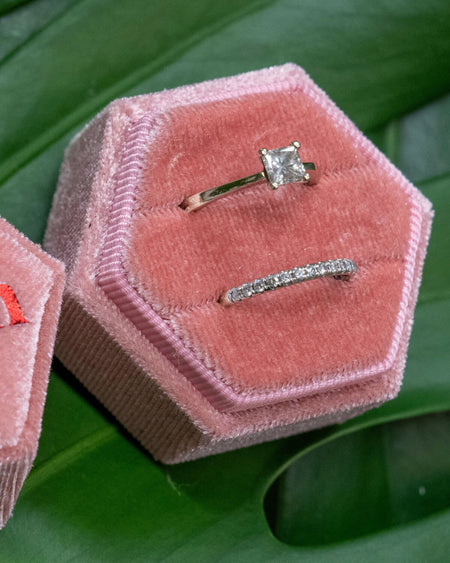 Two open pink hexagonal velvet ring boxes on a green monstera leaf, one showing “’Til Death” embroidered in red and the other holding two elegant silver rings.