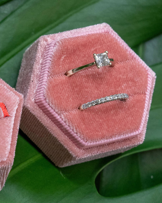 Two open pink hexagonal velvet ring boxes on a green monstera leaf, one showing “’Til Death” embroidered in red and the other holding two elegant silver rings.