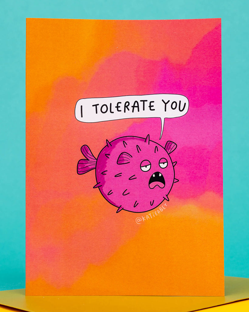 A colourful A6 greeting card from Rebel Romance, designed by Katie Abey, featuring a grumpy purple pufferfish with a speech bubble that says “I tolerate you.” The background is a bold pink and orange gradient. Perfect for sarcastic friendships and alternative love languages. Ideal for millennials who prefer humour over mushy sentiment.