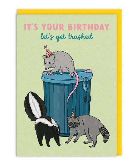 Funny illustrated birthday card from Rebel Romance featuring a possum, raccoon and skunk wearing party hats next to a blue bin. Caption reads "It's Your Birthday, let's get trashed." Set against a pale green background with a bright yellow envelope. Ideal for alternative animal lovers or anyone who parties like a trash panda.