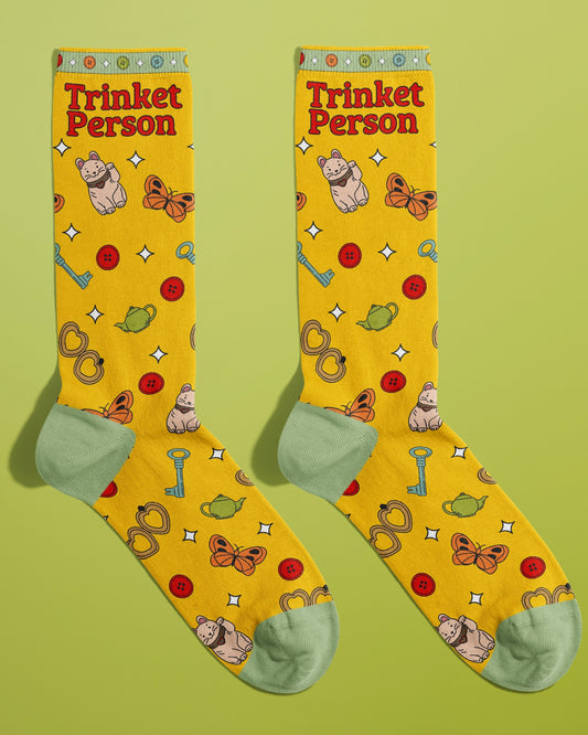 A full view of the Rebel Romance Trinket Person socks designed by FUN CLUB. These yellow crew socks feature the phrase “Trinket Person” and are covered in cute illustrations including lucky cats, butterflies, teapots, keys, red buttons, pretzels, and white sparkles. The toe and heel areas are mint green, and the cuffs have a colourful button pattern. A playful, millennial-friendly accessory for any alternative wardrobe.