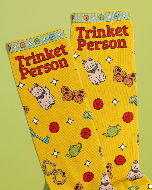 Close-up of Rebel Romance's Trinket Person novelty socks by FUN CLUB, featuring a yellow background with red text reading “Trinket Person.” The design includes lucky cats, butterflies, green teapots, pretzels, keys, red buttons and white sparkle stars. The cuffs are decorated with a row of multicoloured buttons. A fun and quirky fashion accessory perfect for collectors and trinket lovers.