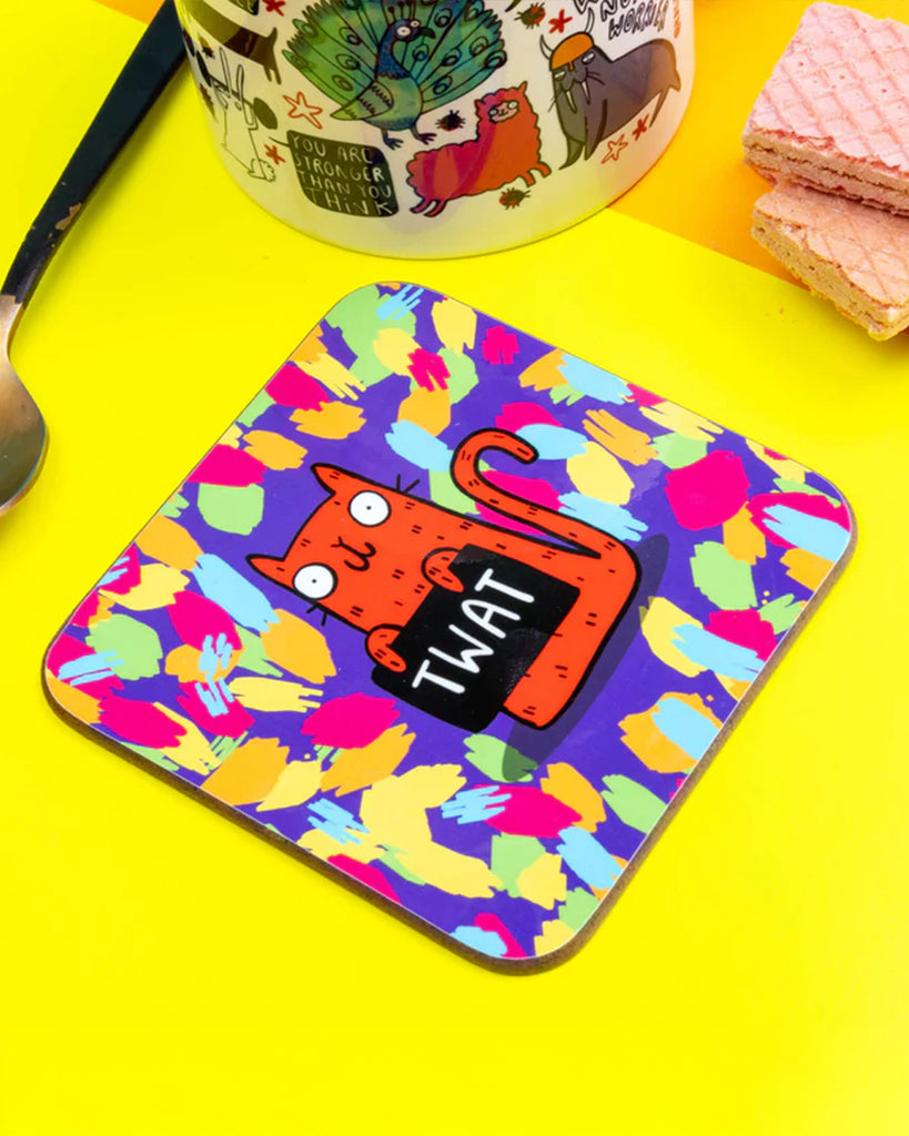 A top-down image of the same coaster lying flat on a yellow surface next to a white illustrated mug and spoon, showcasing the vibrant design and sweary humour.
