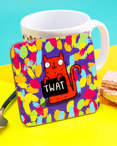 A colourful coaster featuring Katie Abey's red cartoon cat on a multicoloured paint-stroke background, holding a sign that says “TW*T.” The coaster is propped in front of a mug with bright yellow background.