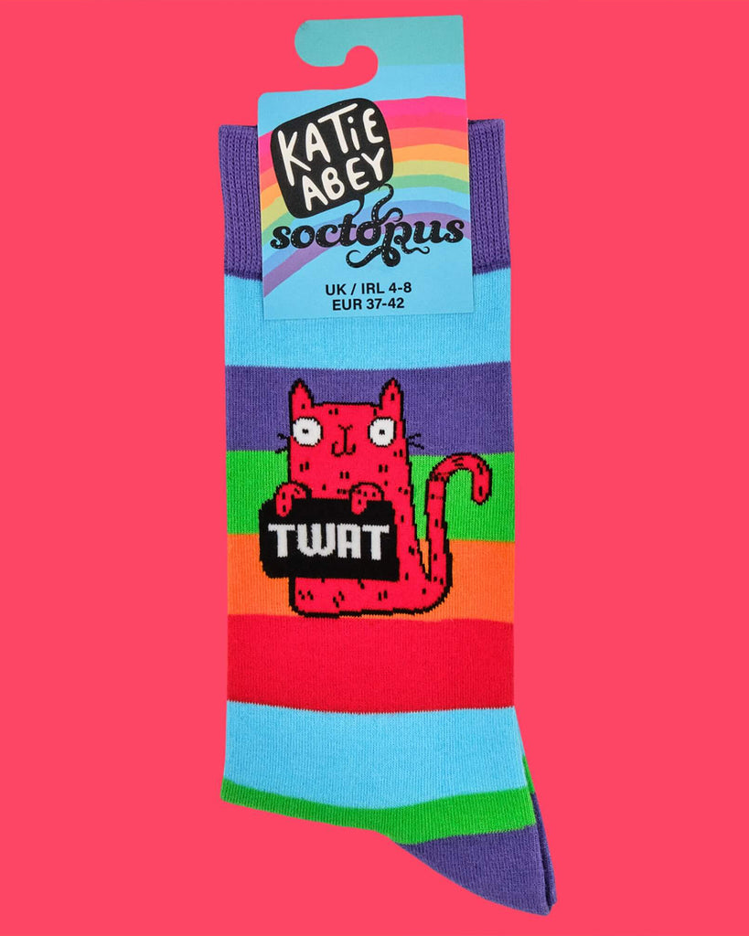 Product shot of a single folded sock against a pink background. The sock shows the full design: a red cat holding a sign reading “TWAT” across rainbow stripes. Label reads “Katie Abey x Soctopus.”