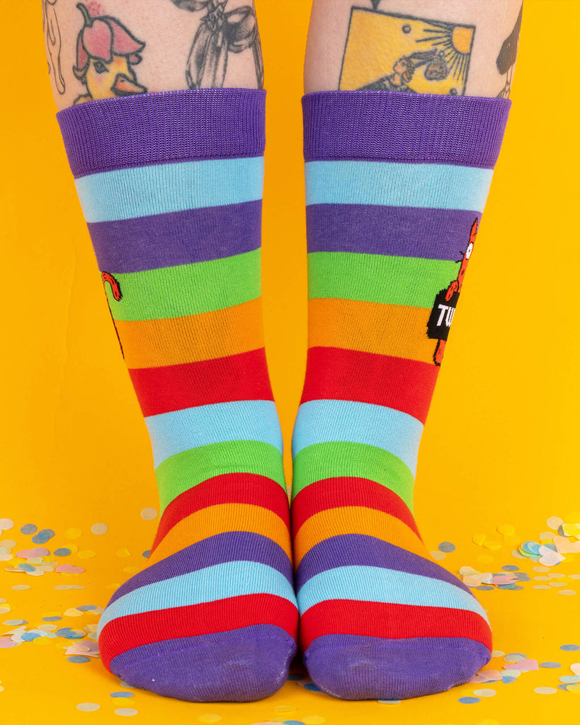 Front view of rainbow-striped Katie Abey socks worn on legs with visible tattoos. The socks feature a red cat holding a sign that says “TWAT” and vibrant rainbow colours from toe to cuff.