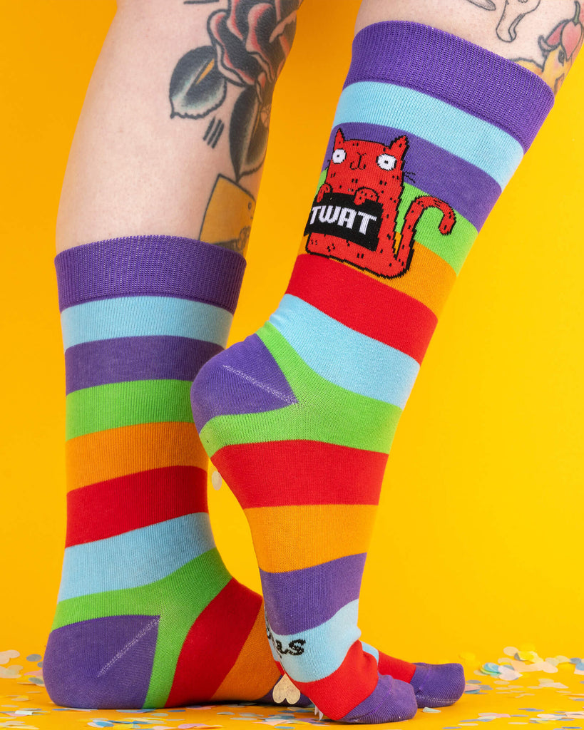 Brightly striped rainbow socks by Katie Abey worn on feet. The design features a red cartoon cat holding a black sign that says “TWAT” in bold white letters. The socks are from the Katie Abey x Soctopus collaboration.