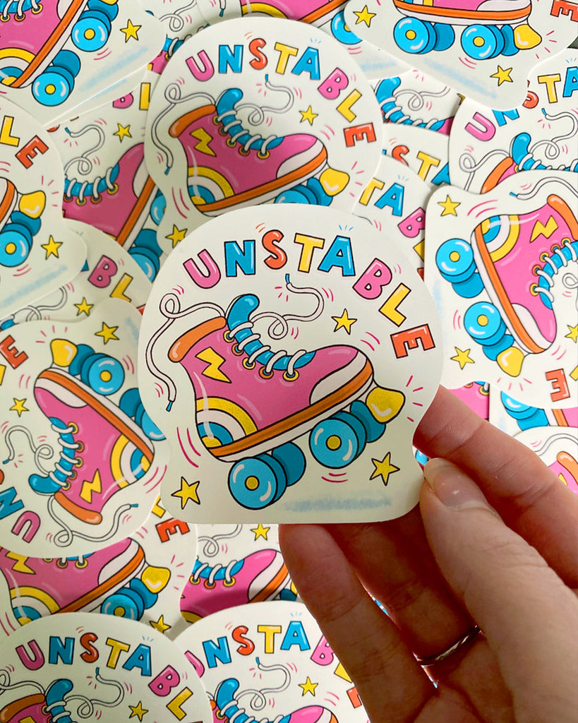 A hand holding a Rebel Romance 'Unstable Rollerskate' sticker against a background filled with the same sticker design. The glossy vinyl sticker shows a pink roller skate with blue wheels, loose laces, and bright lightning bolt details, surrounded by stars and the word "UNSTABLE" in rainbow lettering. Designed by Yasmin Cousins.