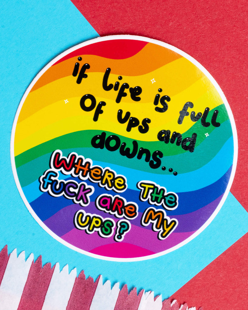 A round rainbow-striped vinyl sticker from Rebel Romance with the bold, cheeky text: “If life is full of ups and downs… where the fuck are my ups?” The design features colourful lettering over a wavy rainbow background. Designed by Innabox, this sticker is waterproof, approx. 8cm wide, and suitable for indoor and outdoor use. Perfect for anyone who needs a dose of humour during hard times.