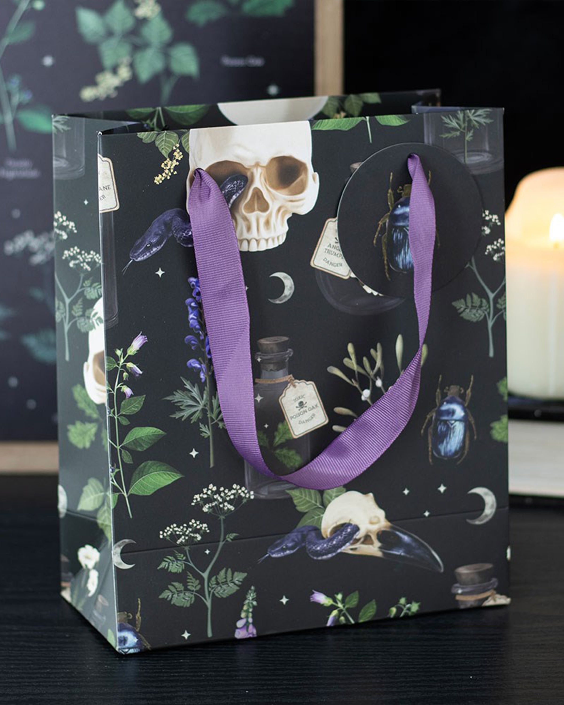 Rebel Romance Medium Venom & Vines Gift Bag styled on a dark surface with a candle in the background, showing its striking skull, snake, and foliage design with purple ribbon handles.