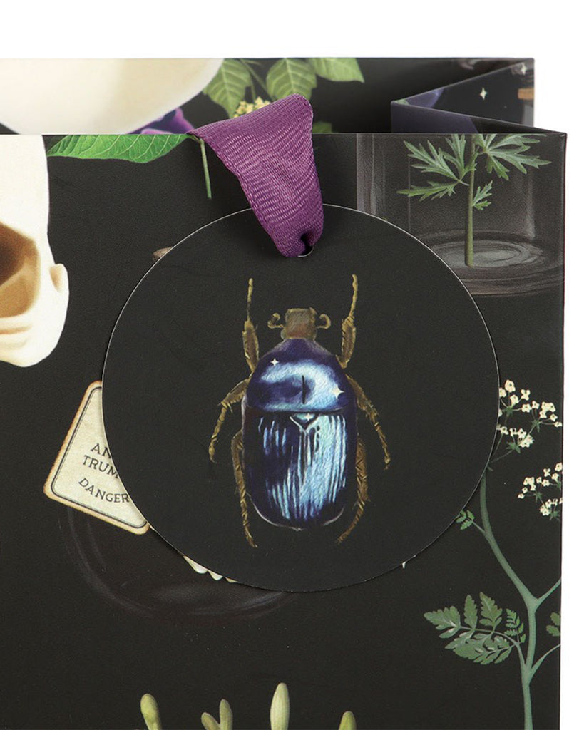 Close‑up of the Rebel Romance Medium Venom & Vines Gift Bag tag, showing a shiny blue beetle on a dark background with botanical details, attached by a purple ribbon handle.