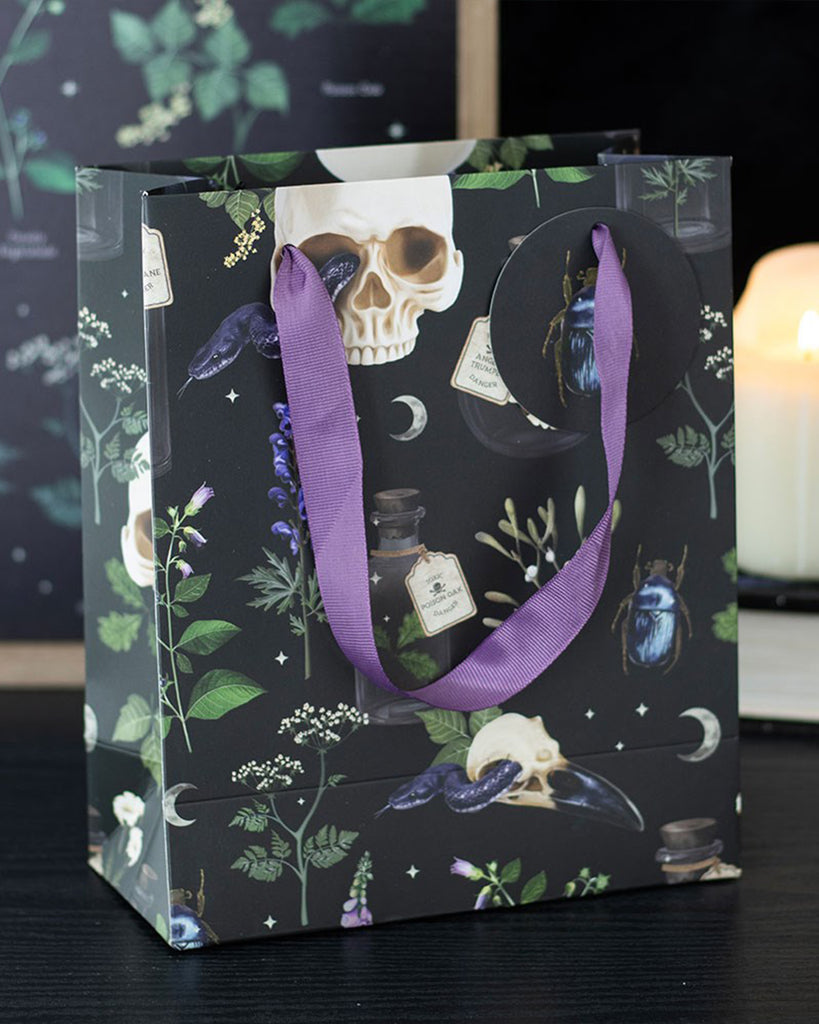 Rebel Romance Medium Venom & Vines Gift Bag styled on a dark surface with a candle in the background, showing its striking skull, snake, and foliage design with purple ribbon handles.