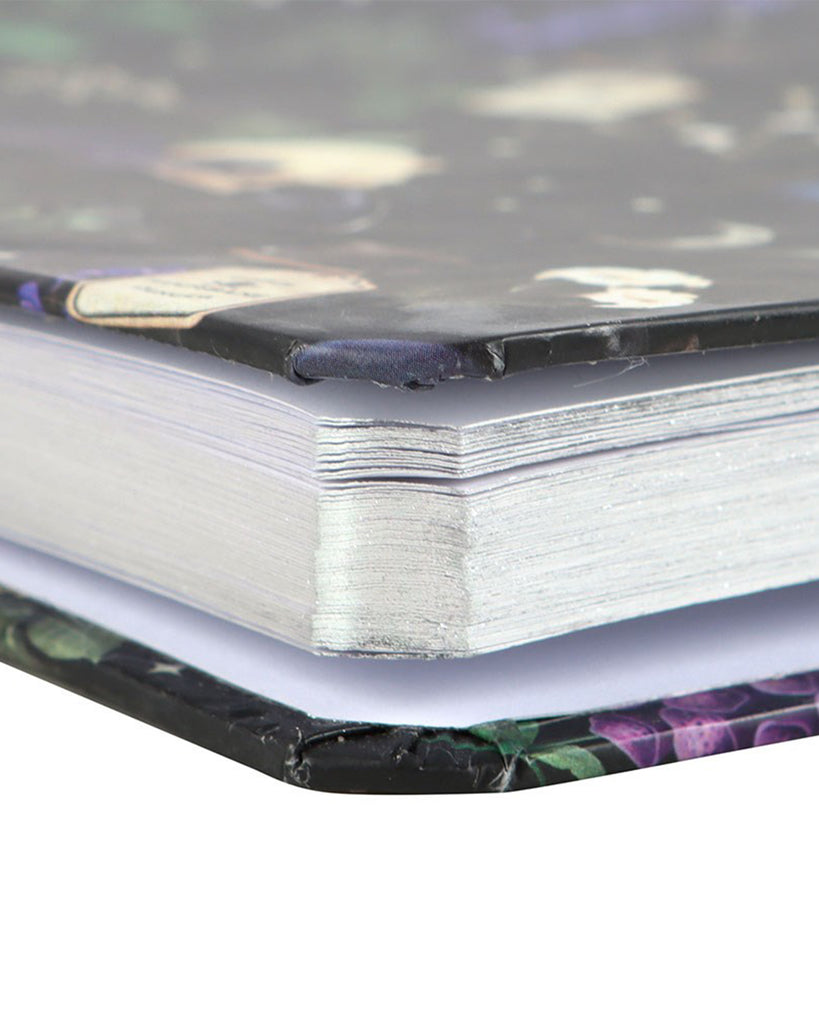 Close-up side view of the Venom & Vines Print A5 Notebook from Rebel Romance, showing the silver-edged lined pages and the glossy hardcover featuring gothic illustrations of skulls, snakes, potions, and poisonous plants.