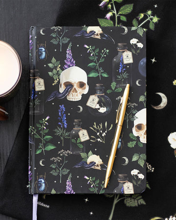 Flat lay of the Venom & Vines Print A5 Notebook from Rebel Romance styled with a gold pen, black celestial-themed cloth, and candle. The notebook features gothic illustrations of skulls, snakes, and herbs on a black background.