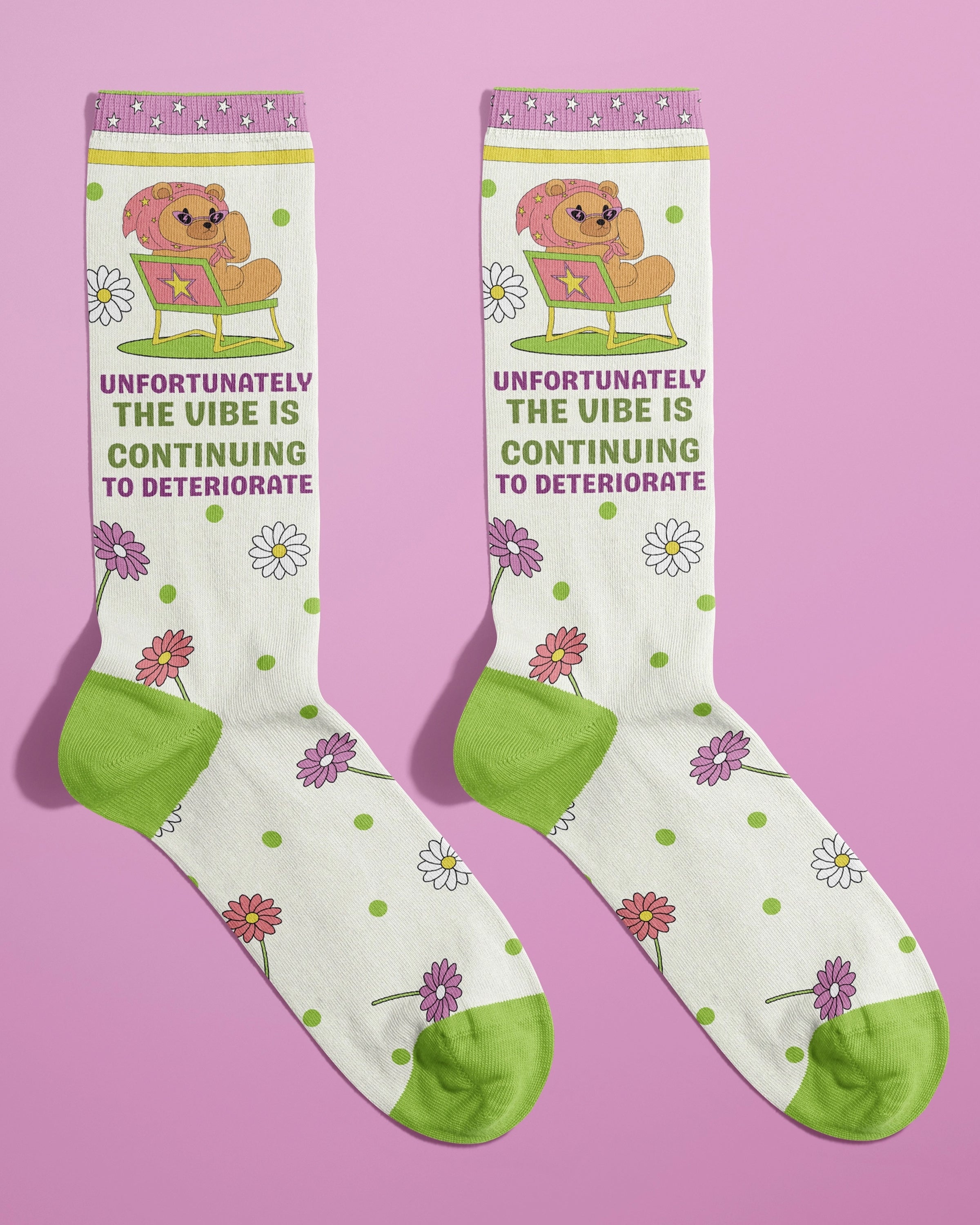 Pair of novelty socks from Rebel Romance shown flat on a pink backdrop. The design includes a lounging bear in heart sunglasses with the phrase “Unfortunately the vibe is continuing to deteriorate” in pink and green text. The socks are decorated with daisies and green polka dots, featuring green toes and heels. Designed by FUN CLUB for alternative UK fashion brand Rebel Romance.