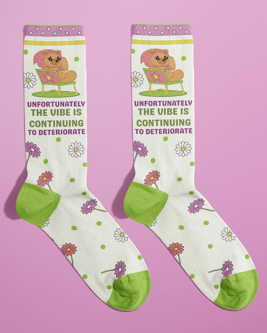 Pair of novelty socks from Rebel Romance shown flat on a pink backdrop. The design includes a lounging bear in heart sunglasses with the phrase “Unfortunately the vibe is continuing to deteriorate” in pink and green text. The socks are decorated with daisies and green polka dots, featuring green toes and heels. Designed by FUN CLUB for alternative UK fashion brand Rebel Romance.
