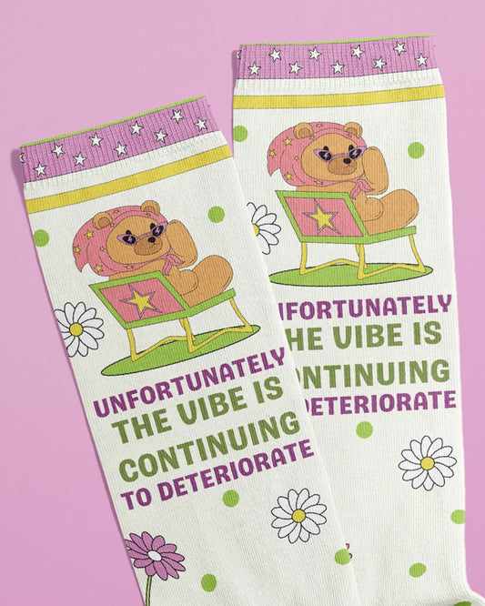 Close-up image of novelty socks by Rebel Romance, featuring a bear in sunglasses and a headscarf lounging on a deck chair. Surrounded by daisies and green polka dots, the socks read “Unfortunately the vibe is continuing to deteriorate” in bold pink and green text. Designed by FUN CLUB. Displayed on a pink background. Funny, alternative style for millennial fashion lovers in the UK.
