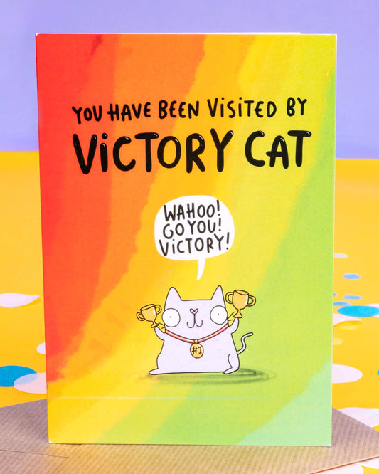 A colourful A6 greeting card from Rebel Romance featuring a cheerful illustrated white cat by Katie Abey holding two gold trophies and wearing a #1 medal. The text reads "You have been visited by Victory Cat" with the cat saying "Wahoo! Go you! Victory!" in a speech bubble. The rainbow background adds a joyful, celebratory vibe. Perfect for congratulating someone on a win or success.